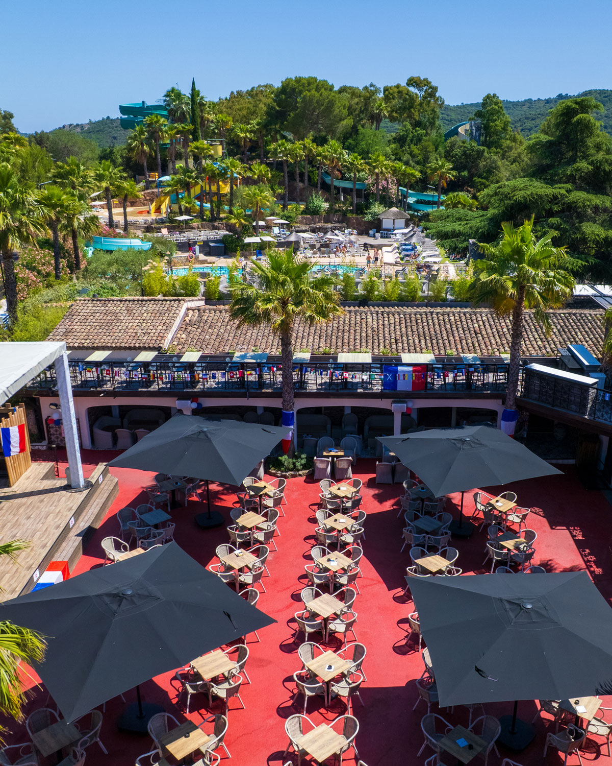 Terras van de bar van Esterel Caravaning onder de parasols, met palmbomen en de glijbanen van het waterpark op de achtergrond